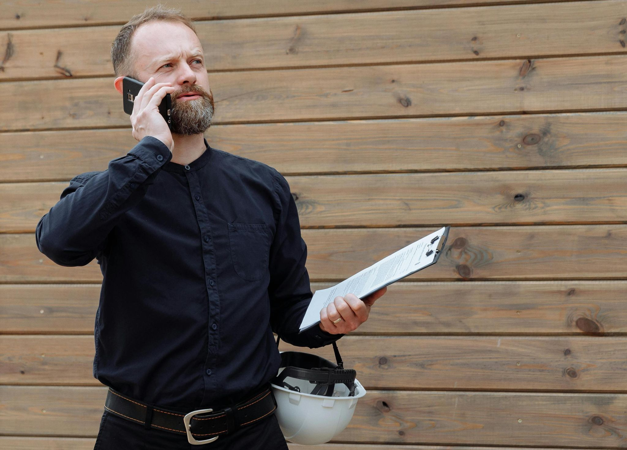 Property manager on the phone outside a home, holding a white hardhat and clipboard